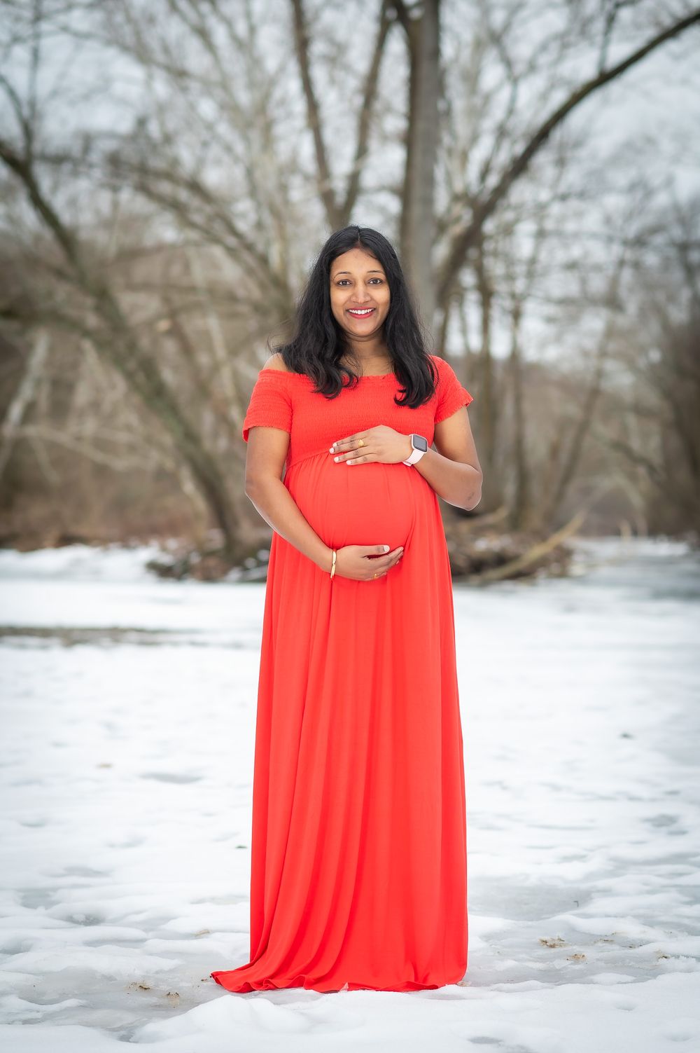 A person in a bright coral dress poses in the snow with bare winter trees in the background.