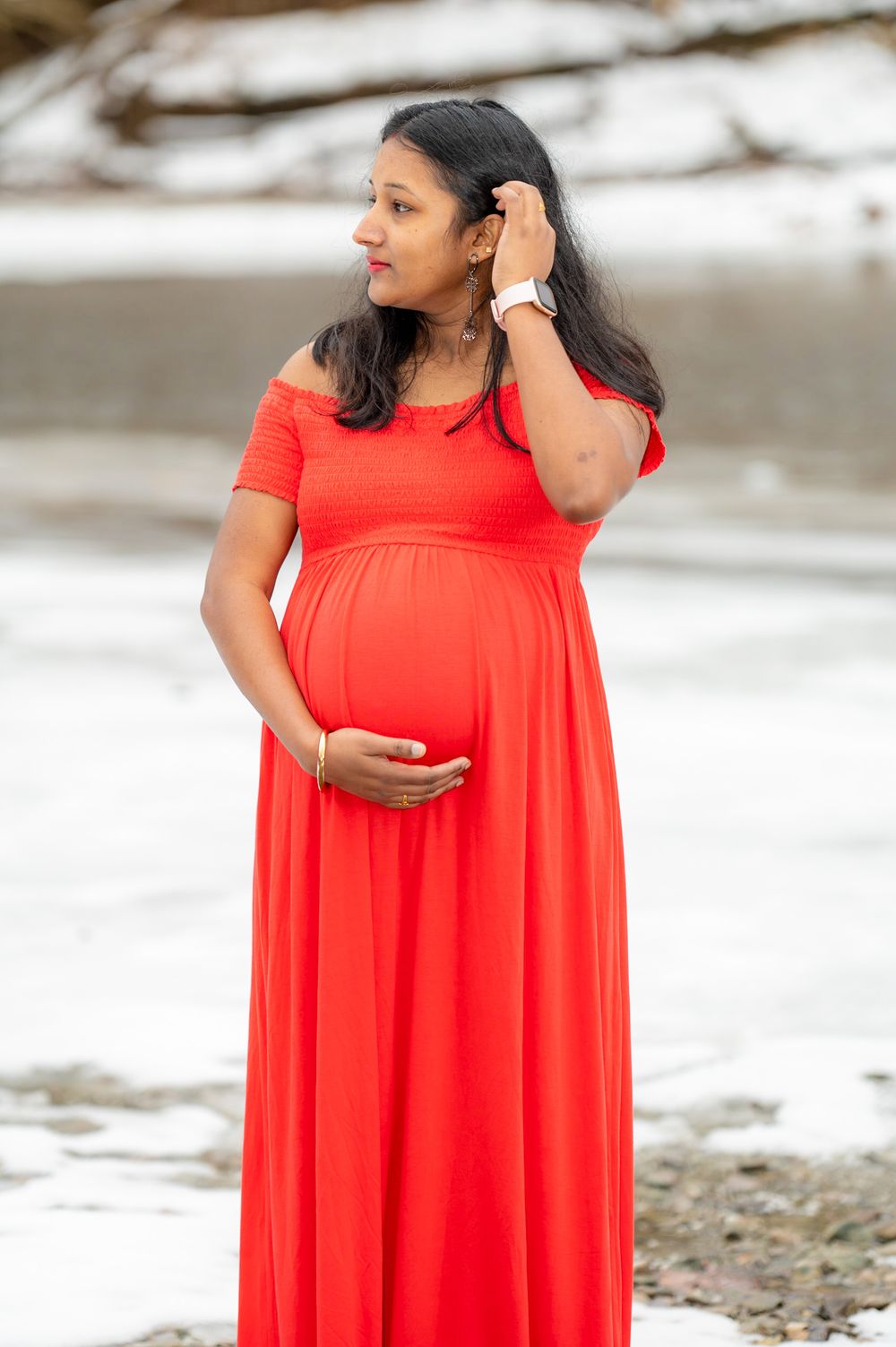 Expectant woman in flowing red maxi dress poses outdoors near snowy landscape while touching her hair.