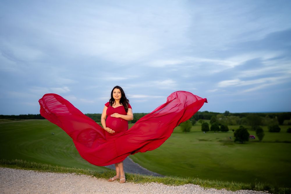 A flowing red dress billows dramatically in the wind against a scenic landscape with blue sky and green fields.