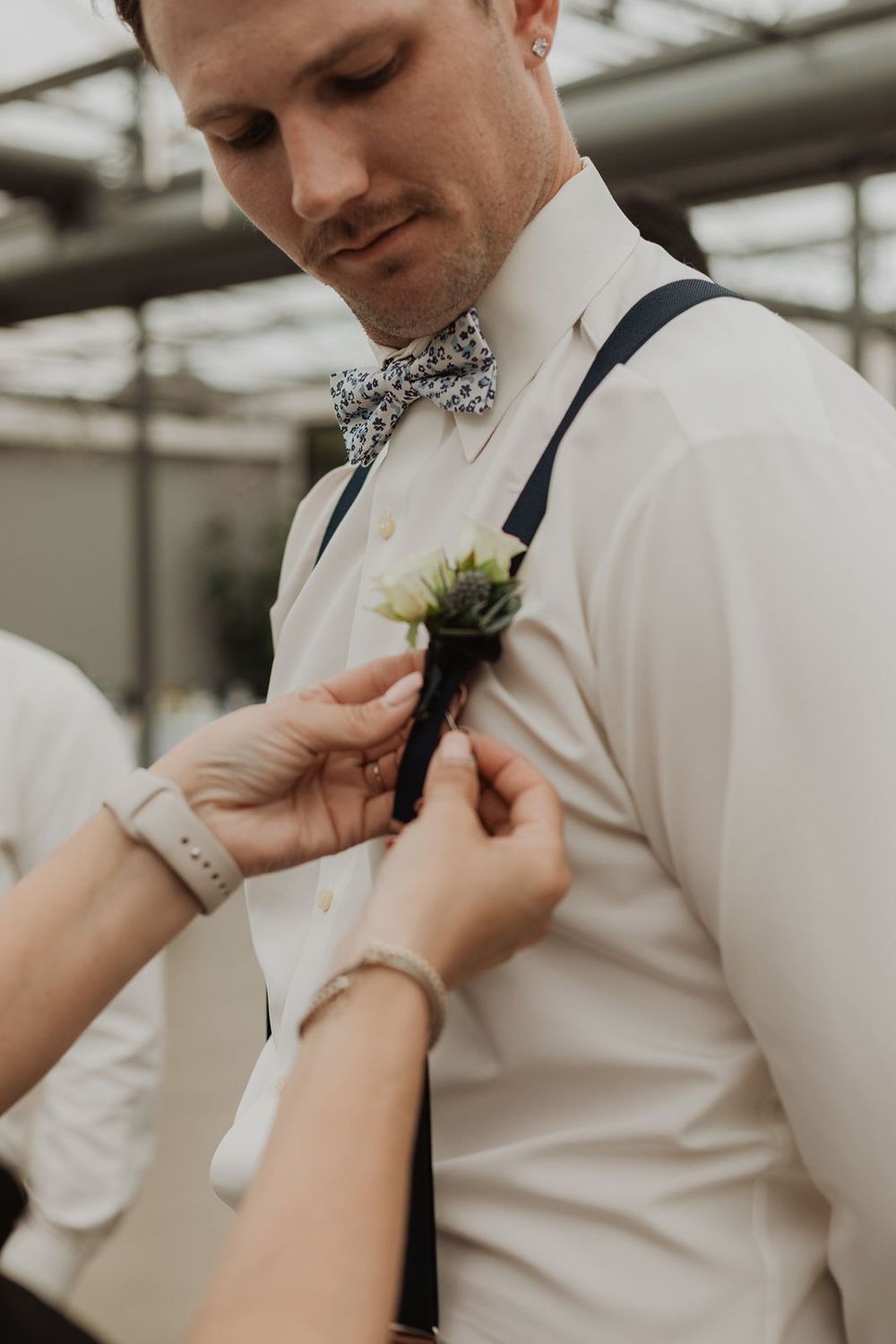 Groomsman in white shirt and navy suspenders having boutonniere pinned on at wedding preparation.