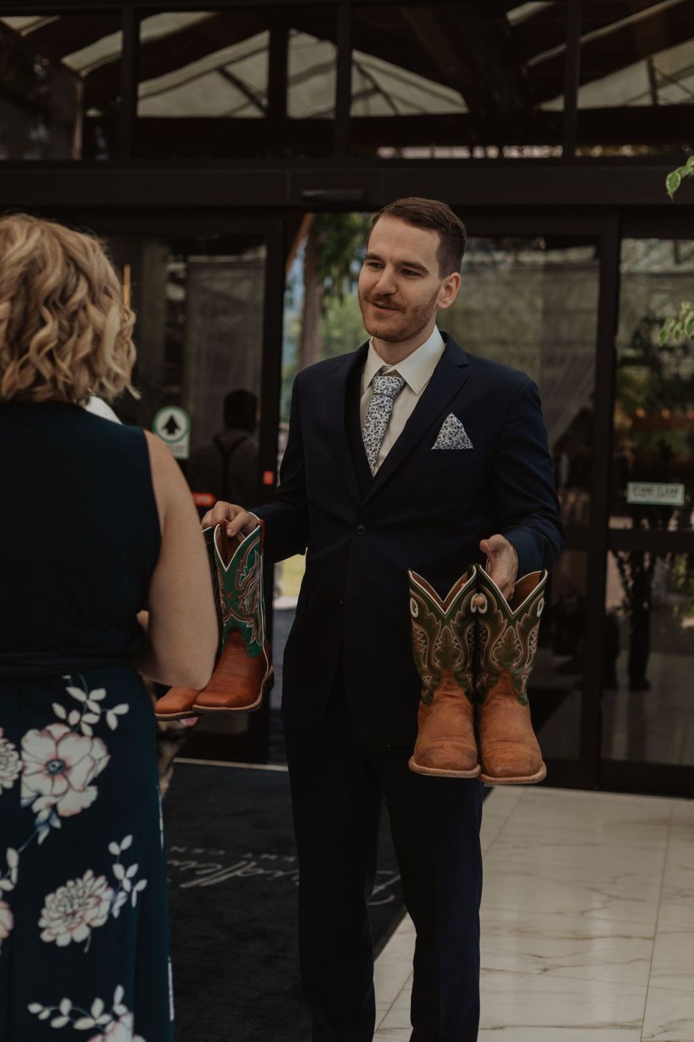 Smiling groom in navy suit holding two pairs of cowboy boots at the wedding venue.