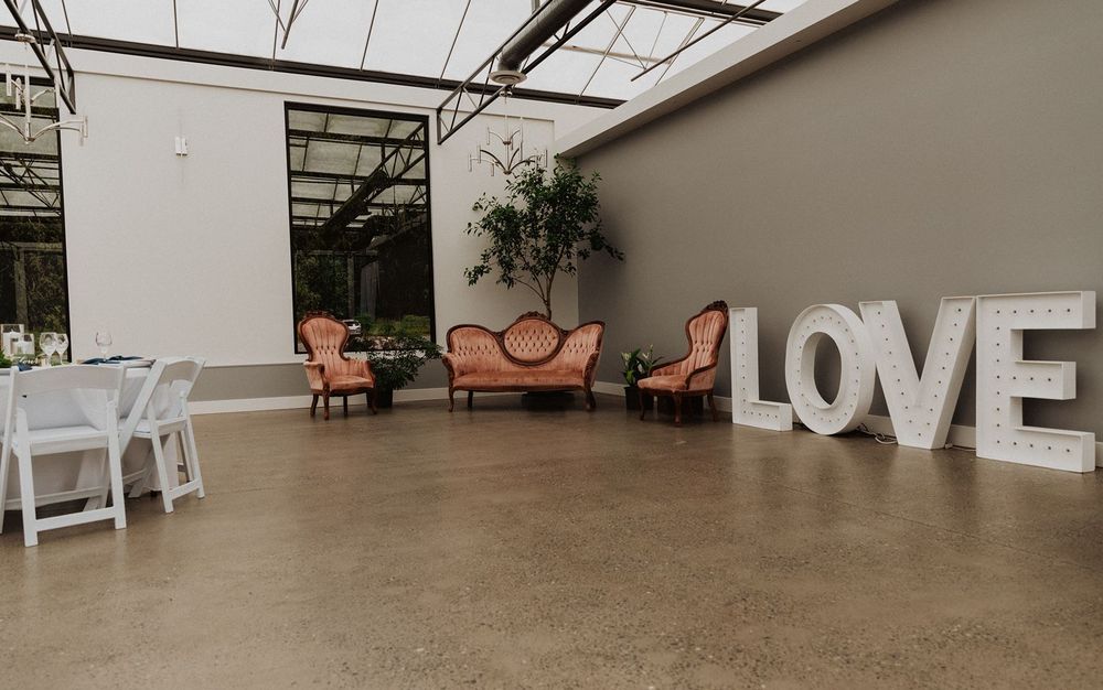 Elegant wedding venue with marquee LOVE letters, vintage pink velvet sofas, and white folding chairs on polished concrete floors.