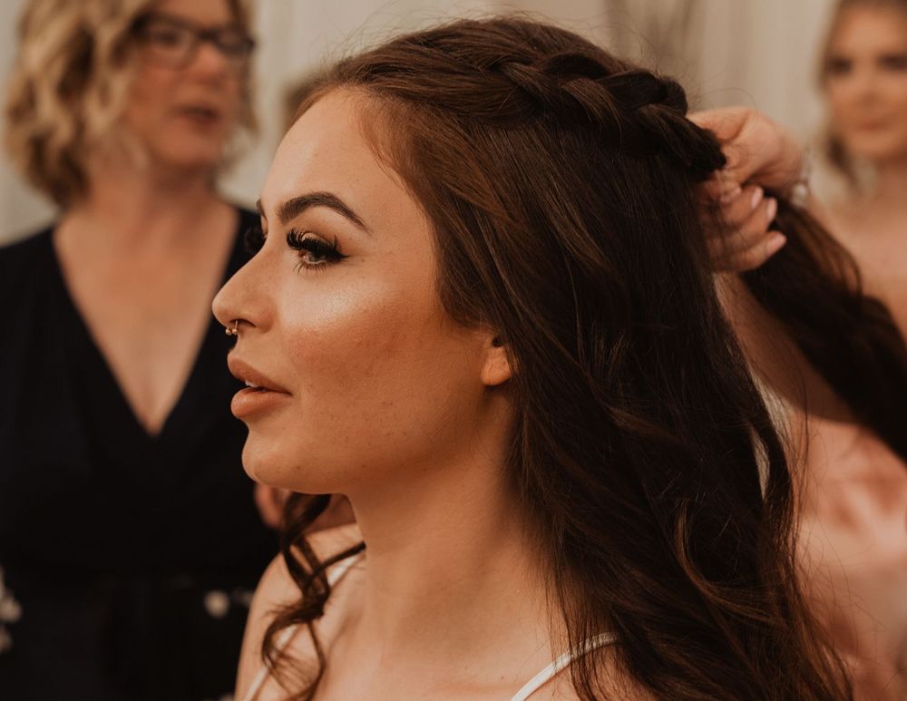 Bride getting her hair styled into a braided half-updo with soft waves by a hairstylist at a wedding.