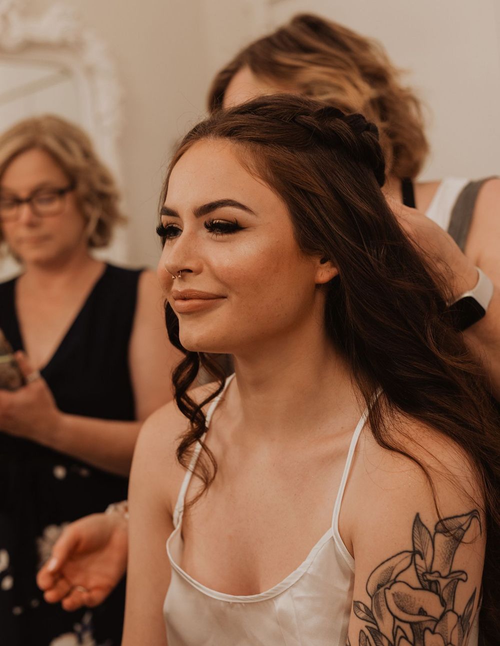 Bride with long dark wavy hair, braided updo, and floral tattoo on shoulder smiling during wedding preparations.