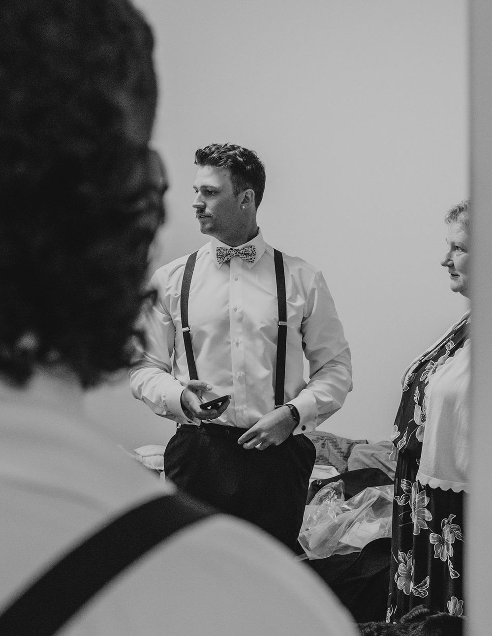 Groom in suspenders and bow tie holding phone at wedding reception in black and white photo.