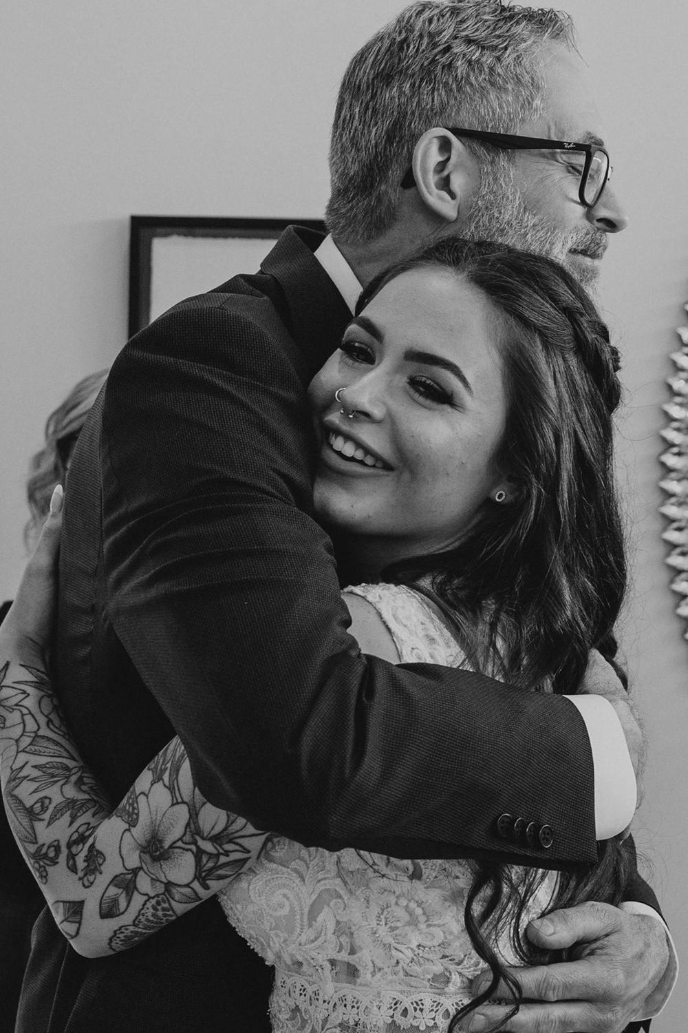 Smiling bride with tattoos hugs older man in suit in black and white wedding photo.