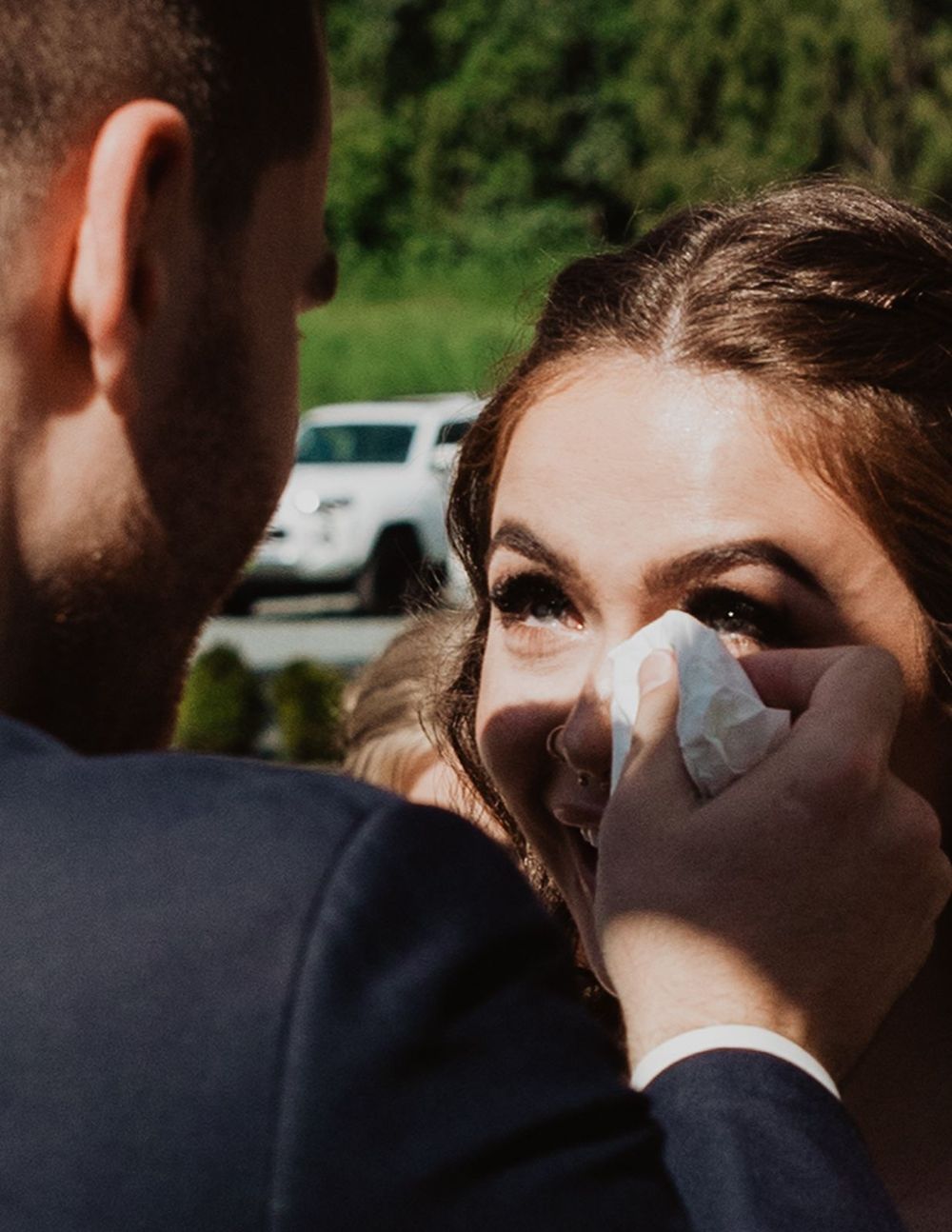 Groom tenderly wipes tears from emotional brides eyes during outdoor wedding ceremony.
