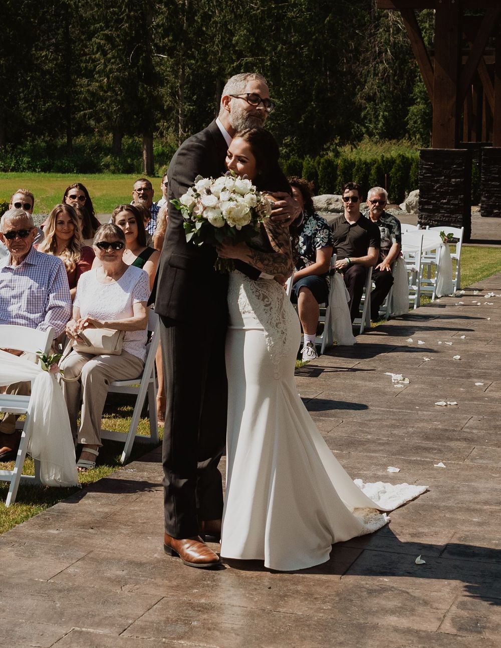 Father kisses bride on head during emotional aisle walk at outdoor summer wedding.