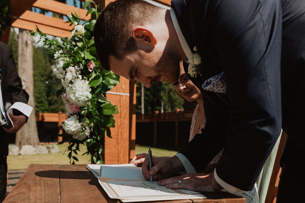 Groom signing wedding certificate at wooden table decorated with floral greenery arch.