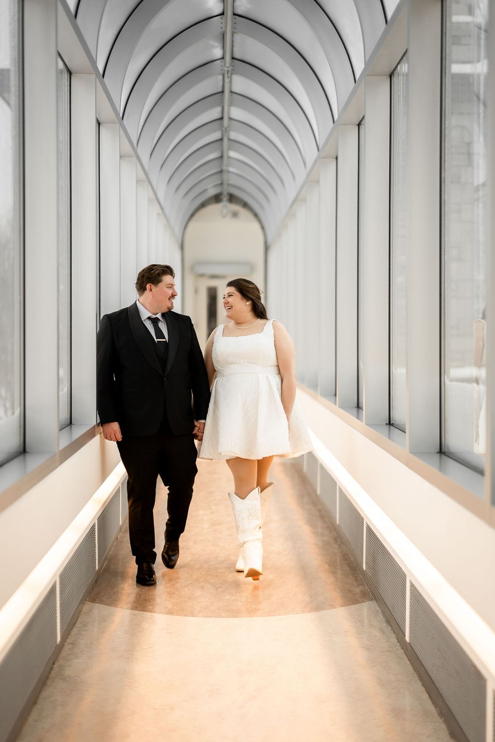 A wedding couple walks through a white arched corridor with dramatic architectural lighting.