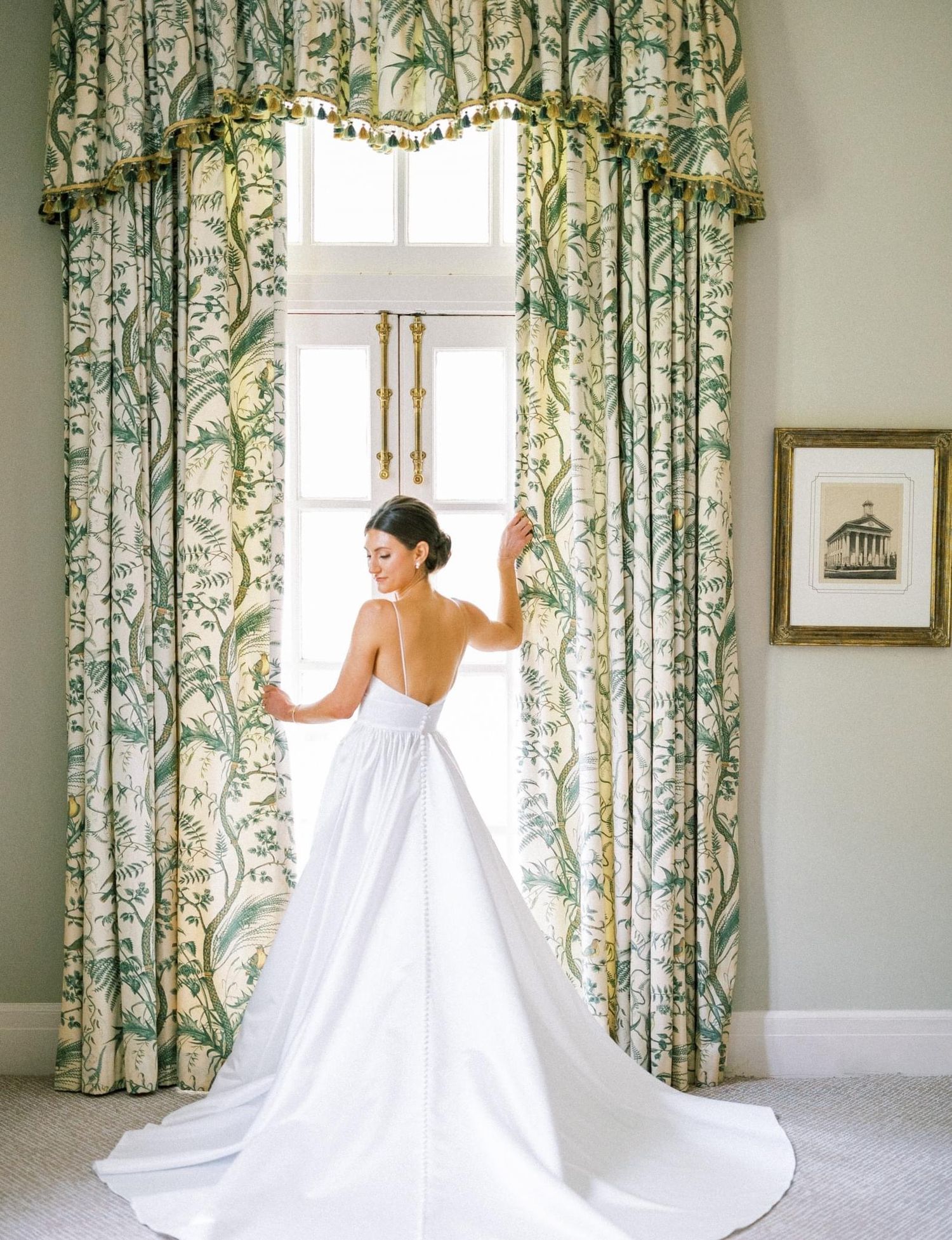 A bride in a flowing white gown poses gracefully in an elegant room with floral curtains and soft natural lighting.