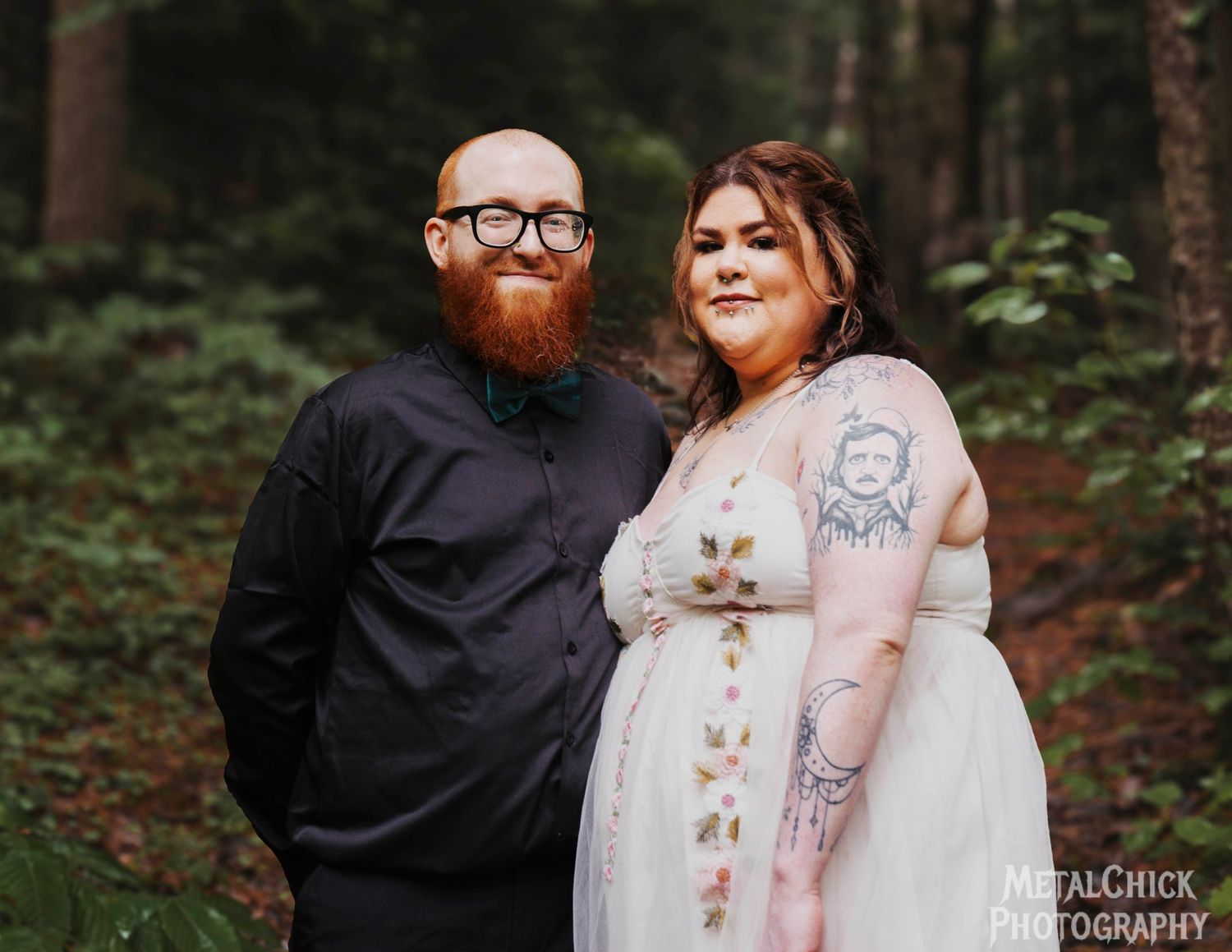 Wedding portrait of a bride with tattoos and a groom with a red beard standing in a lush green forest.