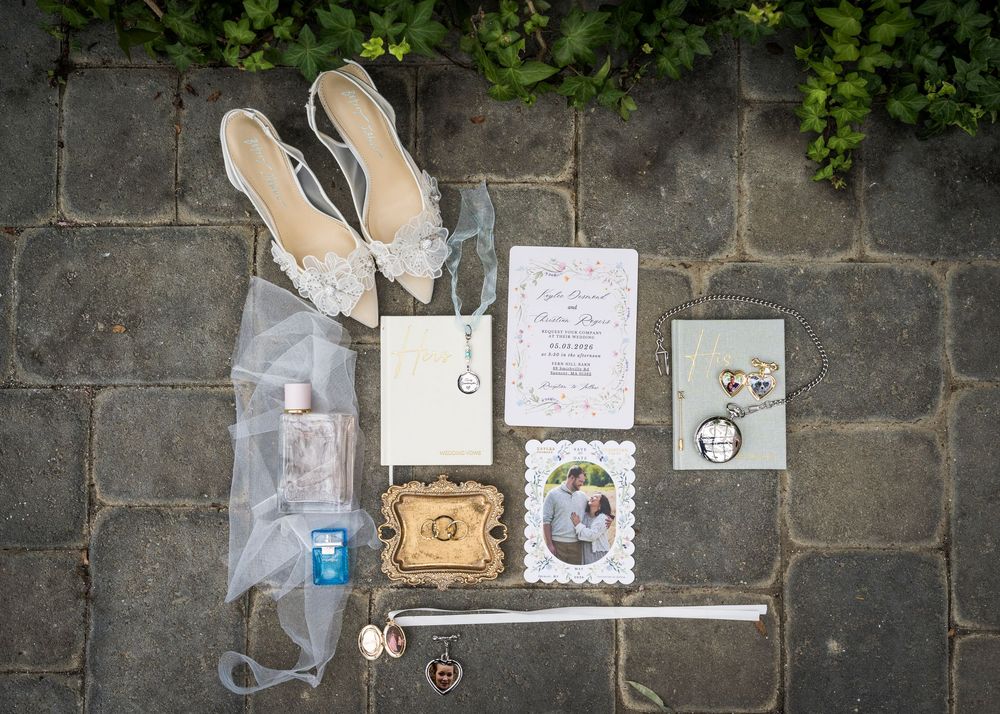 Flat lay of wedding details including lace heels, veil, invitation, photo, and accessories on stone pavement.