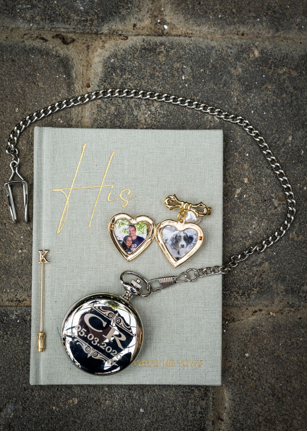 A silver pocket watch with engraved initials rests on a gray journal beside gold heart lockets with photos and a decorative pin.