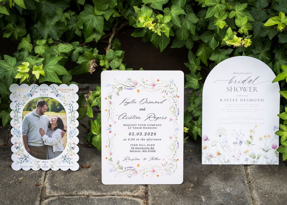 Wedding invitation suite with wildflower design, bridal shower sign, and couple photo displayed against green foliage backdrop.