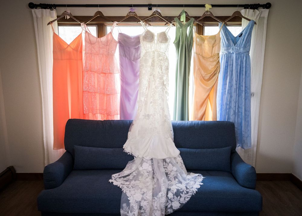White lace wedding gown displayed with colorful bridesmaid dresses in rainbow hues above blue sofa.