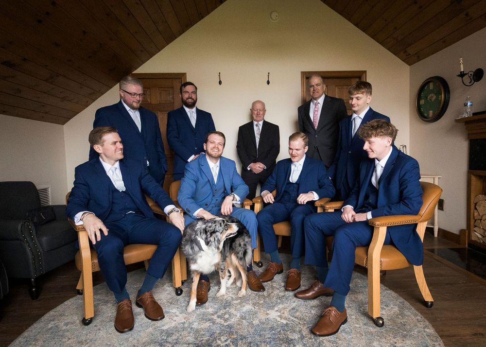 Groomsmen in navy suits posing with a dog in rustic wood-paneled room before wedding ceremony.