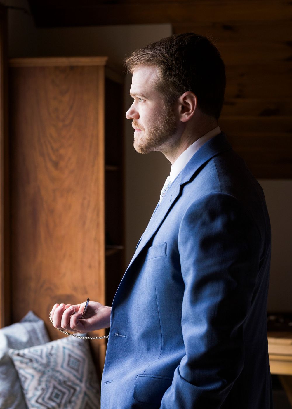 Groom in blue suit holding pocket watch chain by window in soft natural light.