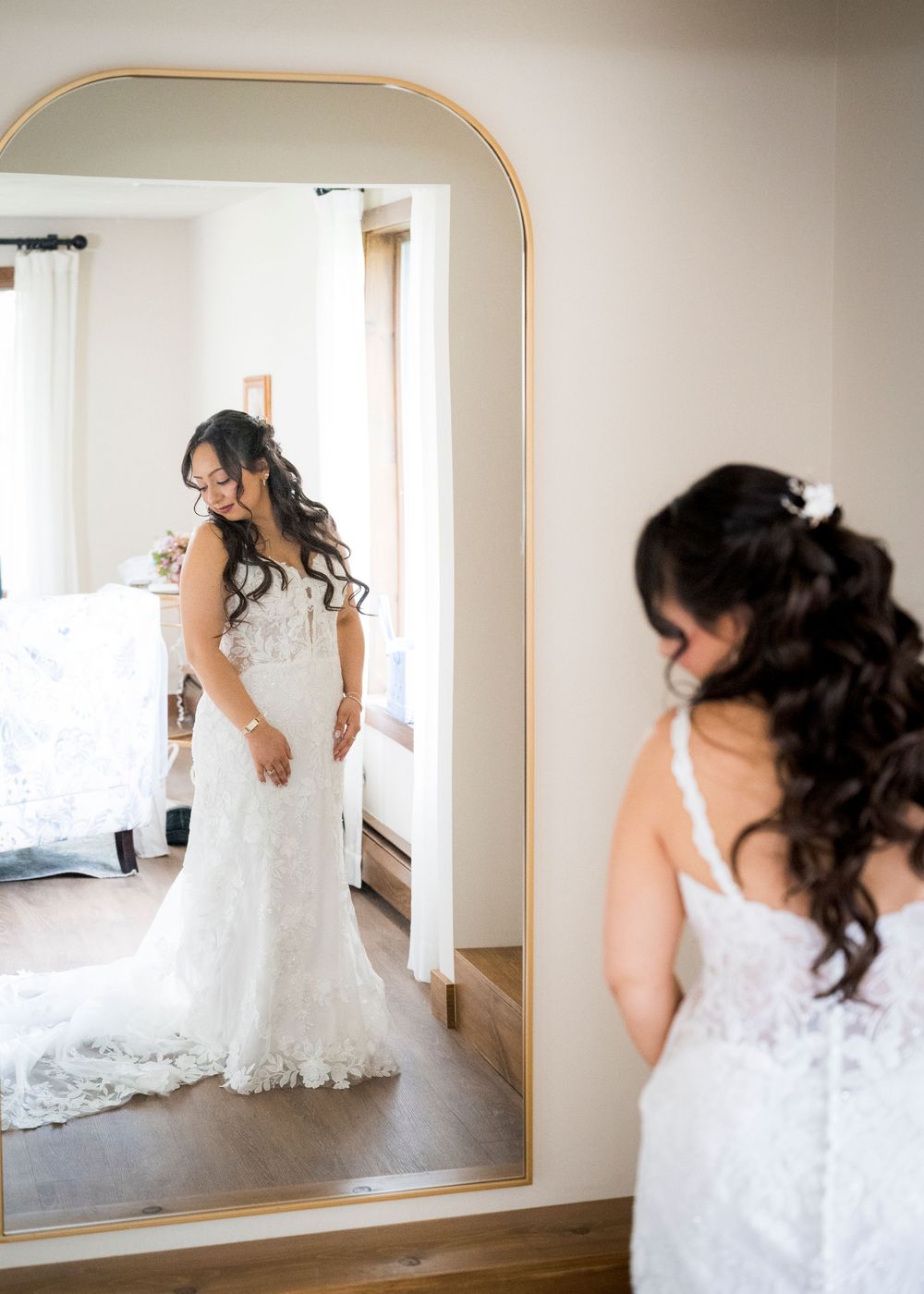 Bride in white lace wedding dress admires her reflection in a large gold-framed floor mirror.
