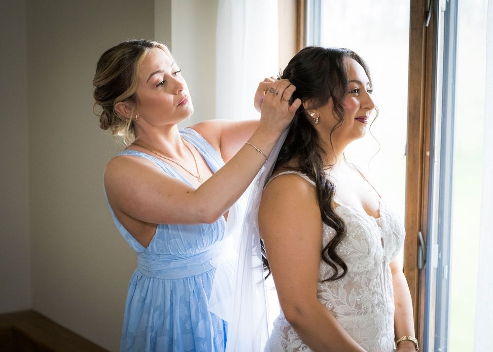Bridesmaid in blue dress adjusting bride's veil near window during wedding day preparations.