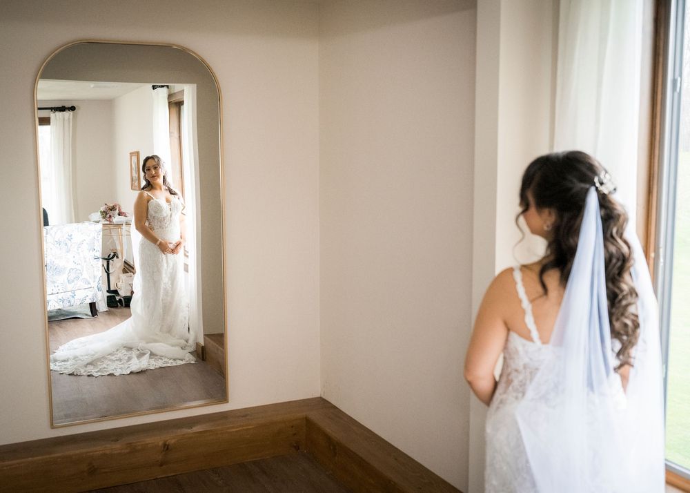 Bride in white lace wedding gown admiring her reflection in an arched mirror before her wedding ceremony.