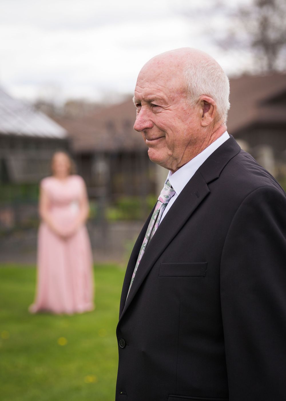 An elderly man in a black suit smiles outdoors with a woman in a pink gown in the background.