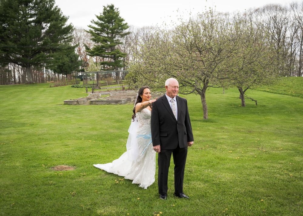 Bride in lace wedding gown tapping father on shoulder for first look on green lawn with spring trees.