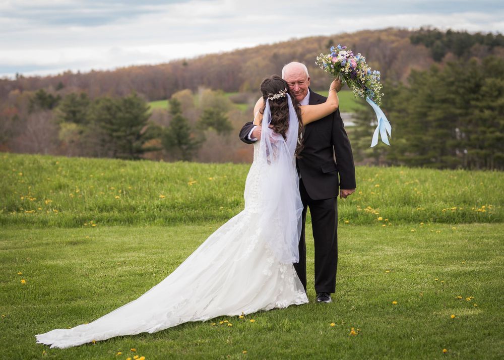 Bride in elegant lace gown embraces older gentleman on lush green lawn with wooded hills behind.