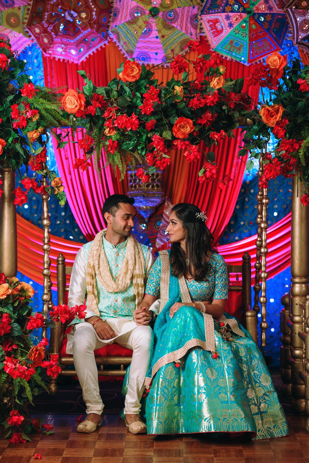 Couple in matching turquoise Indian wedding attire seated on ornate stage with red floral decorations and pink drapes.