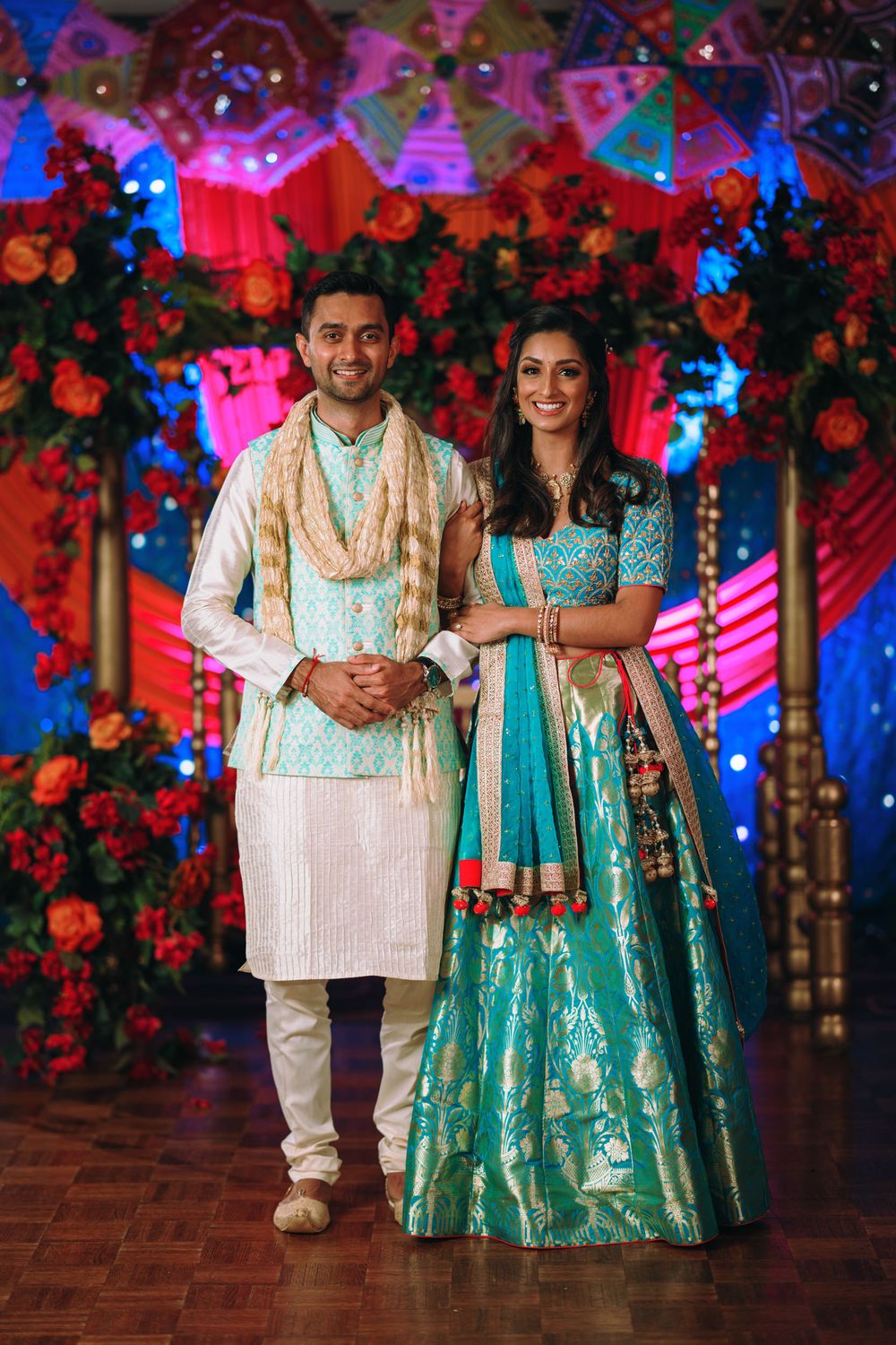 A couple in traditional Indian attire poses at a colorful wedding venue decorated with red flowers and blue lighting.
