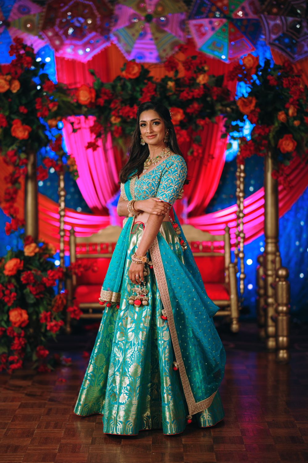 A person in a stunning turquoise and gold lehenga poses against a vibrant red and blue decorated backdrop.