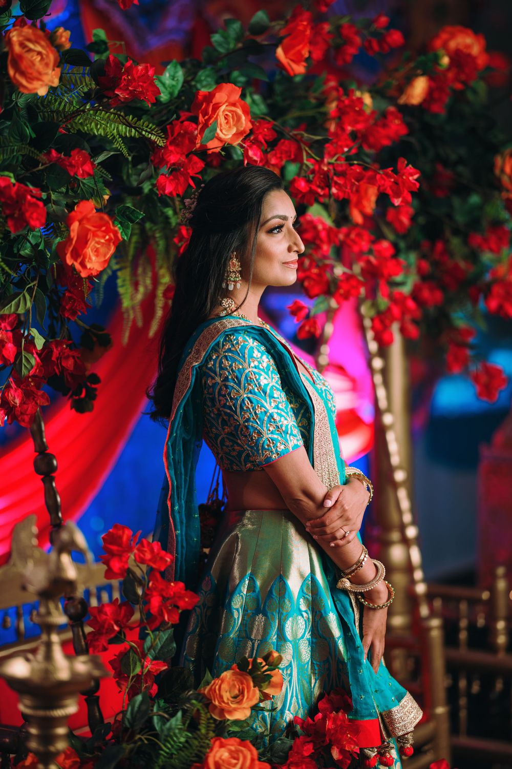 Traditional Indian bride in turquoise lehenga poses elegantly against vibrant red floral backdrop with blue lighting.