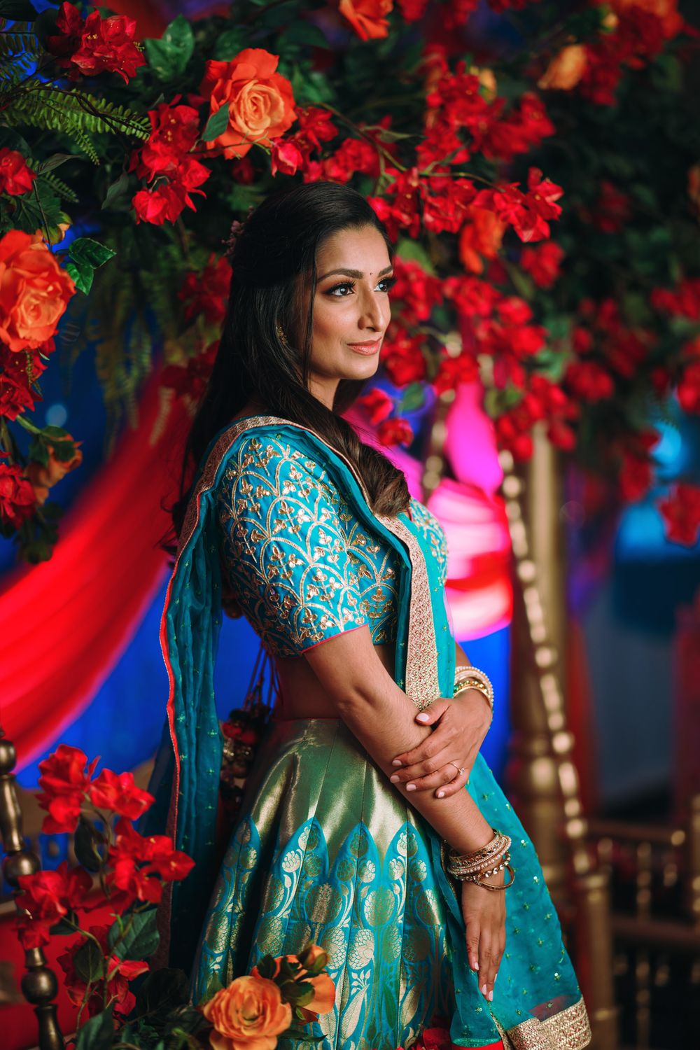 A stunning portrait in a teal and gold embroidered dress against vibrant red floral decorations and blue lighting.