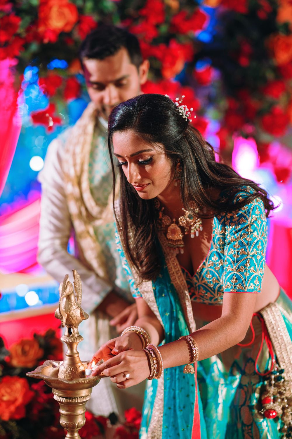 Traditional Indian wedding ceremony detail showing person lighting ceremonial oil lamp in turquoise attire with red decor.