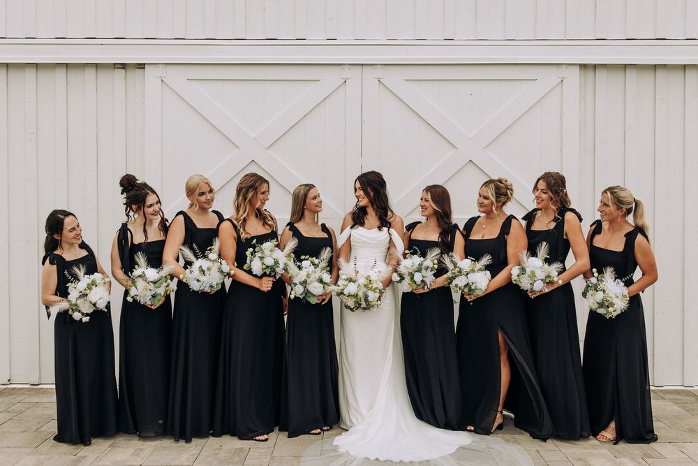 Bride in white gown poses with nine bridesmaids in black dresses holding white bouquets by barn doors.