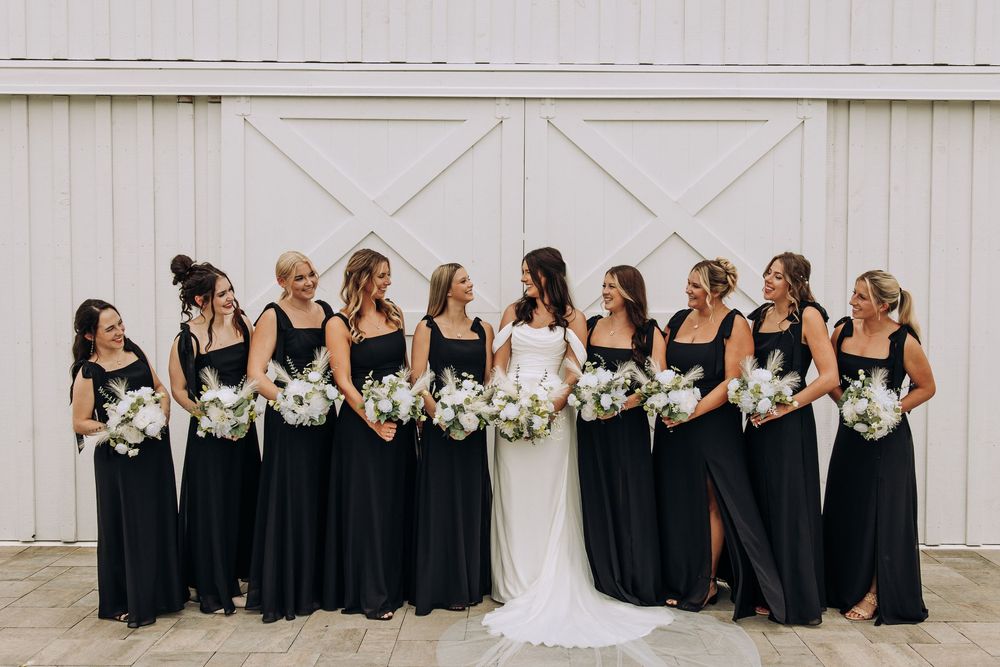 Bride surrounded by laughing bridesmaids in black gowns holding white floral bouquets outside white barn.