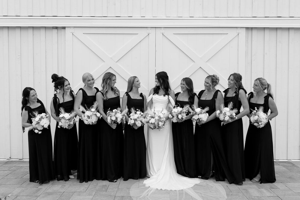 Black and white photo of bride and nine bridesmaids in black dresses laughing outside white barn doors.