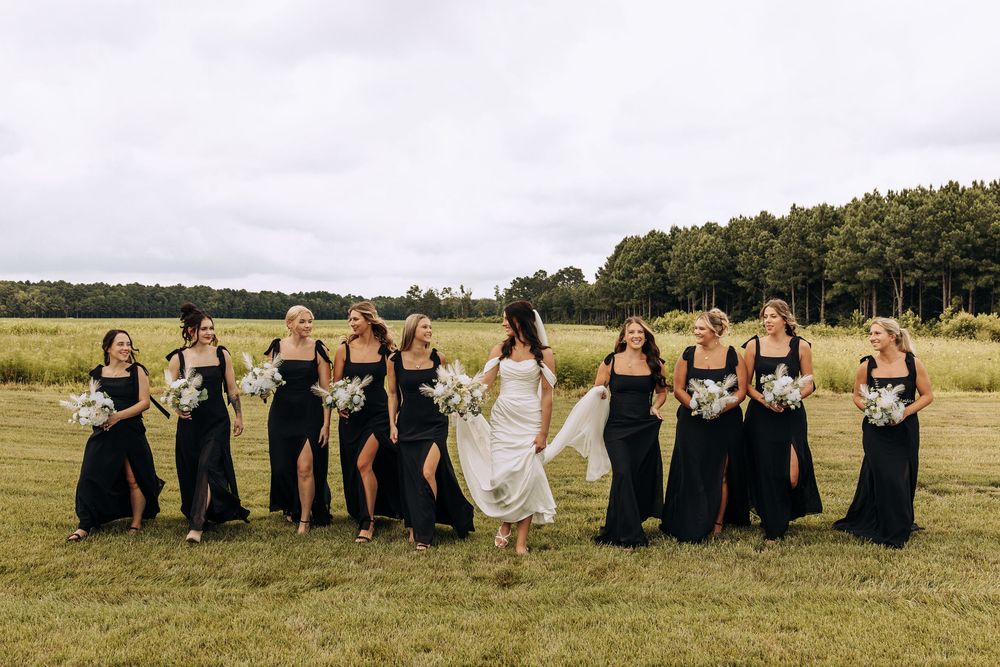 Bride in flowing white dress walks with bridesmaids in black gowns across green field near tree line.