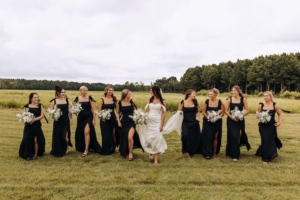 Bride in white gown walks with nine bridesmaids in black dresses holding white bouquets across a green field.