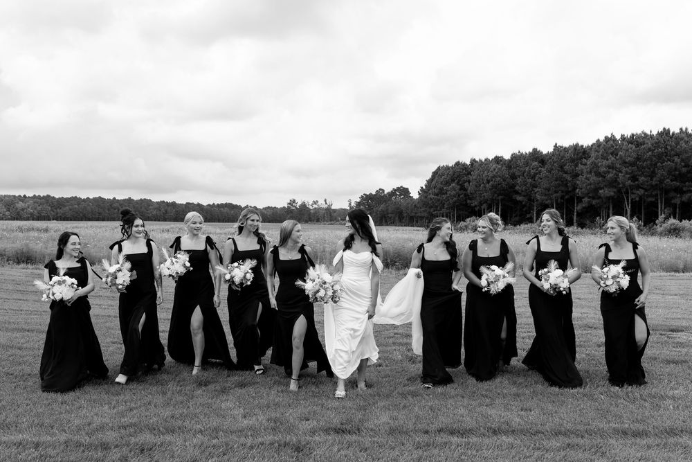 Black and white photo of bride and bridesmaids in black gowns walking through a grassy outdoor venue.