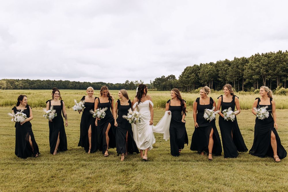 Bride in white dress laughs with bridesmaids in black gowns carrying white floral bouquets on a lawn.