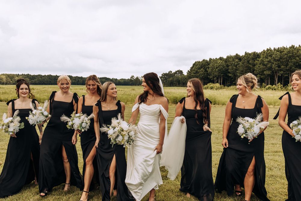 Bride in off-shoulder white gown strolls with smiling bridesmaids in black dresses across an open field.