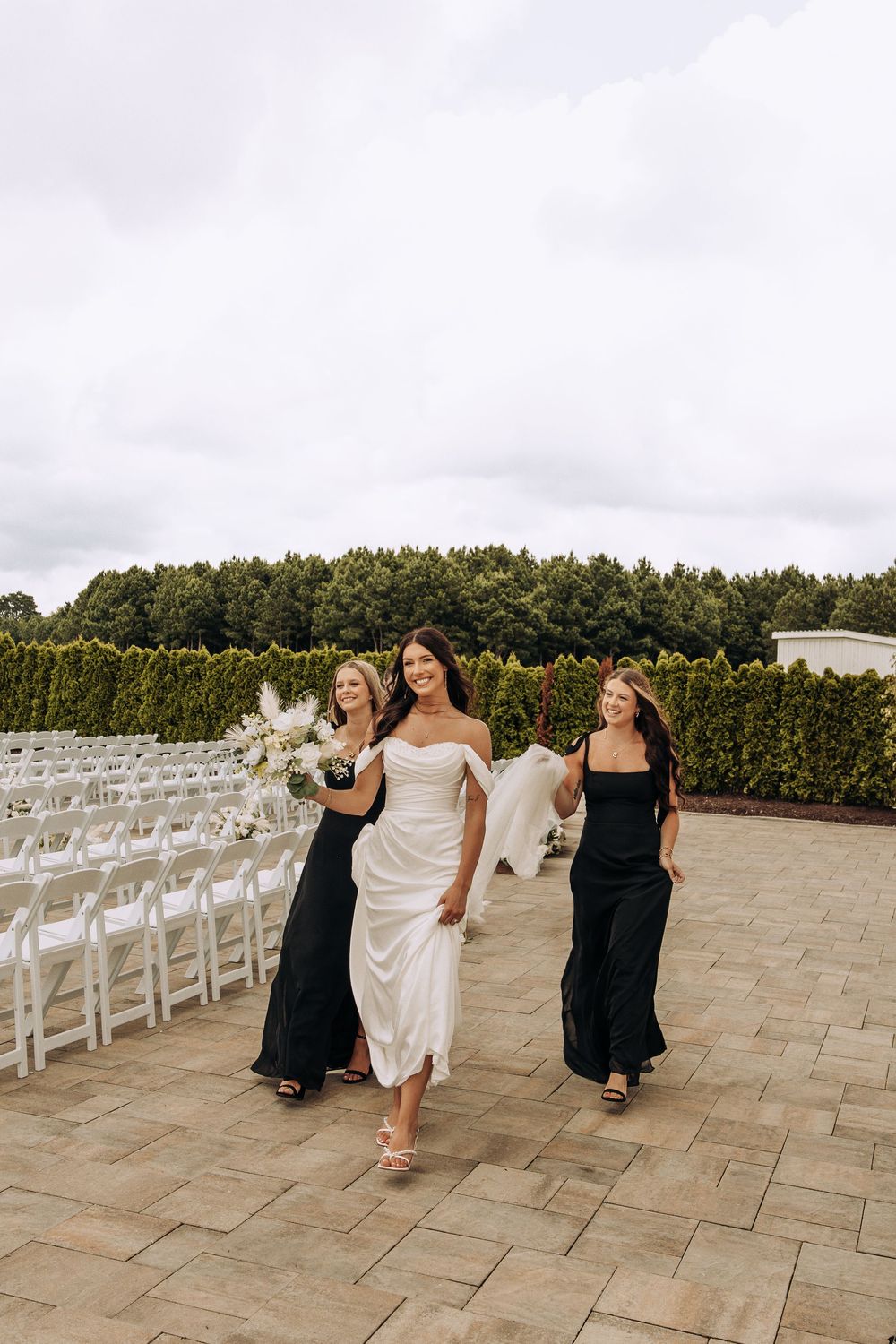 Bride in white satin gown walks with two bridesmaids in black dresses past rows of white ceremony chairs.
