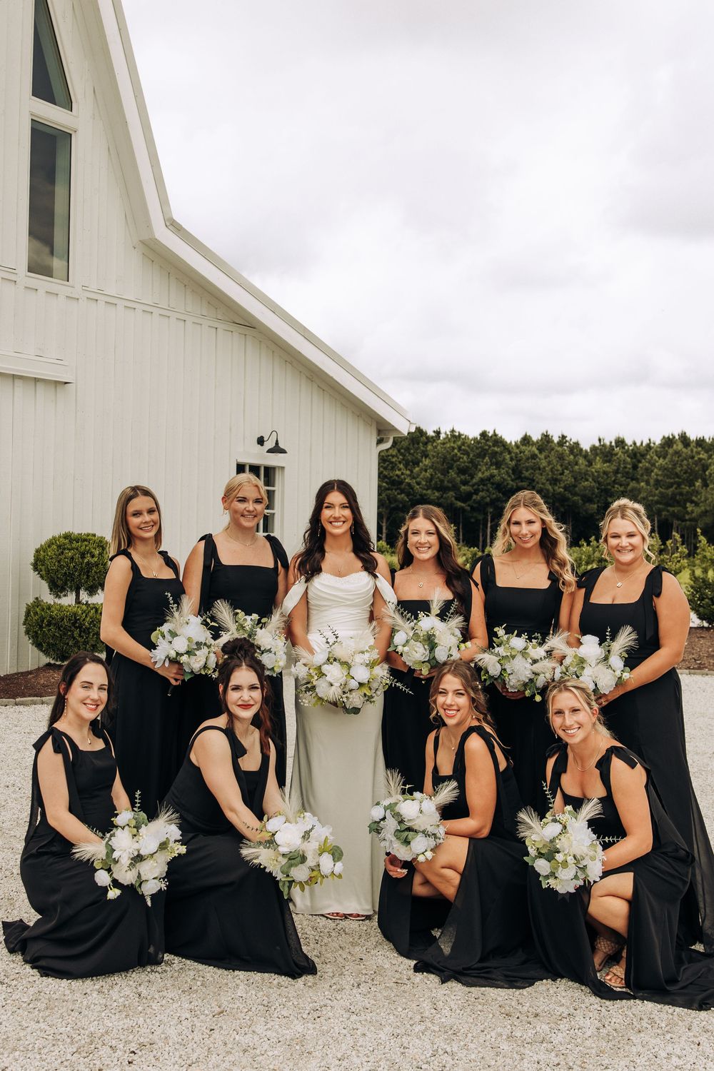 Bride poses with nine bridesmaids in black gowns holding white bouquets outside a white barn venue.
