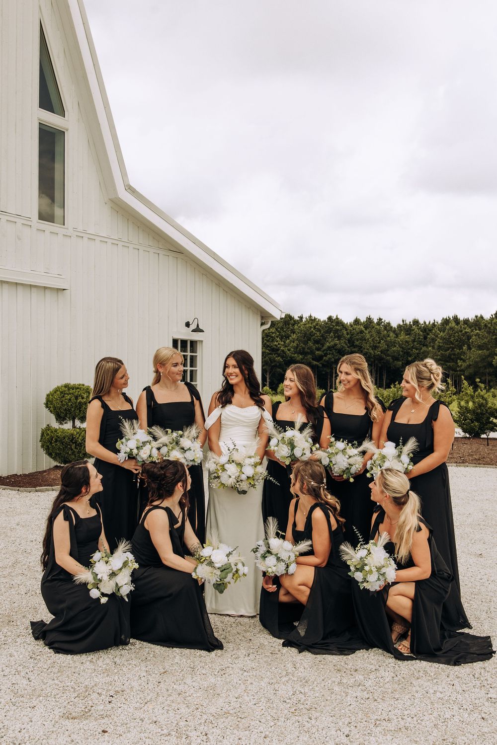 Bride and bridesmaids in black dresses pose with white floral bouquets beside a white barn building.
