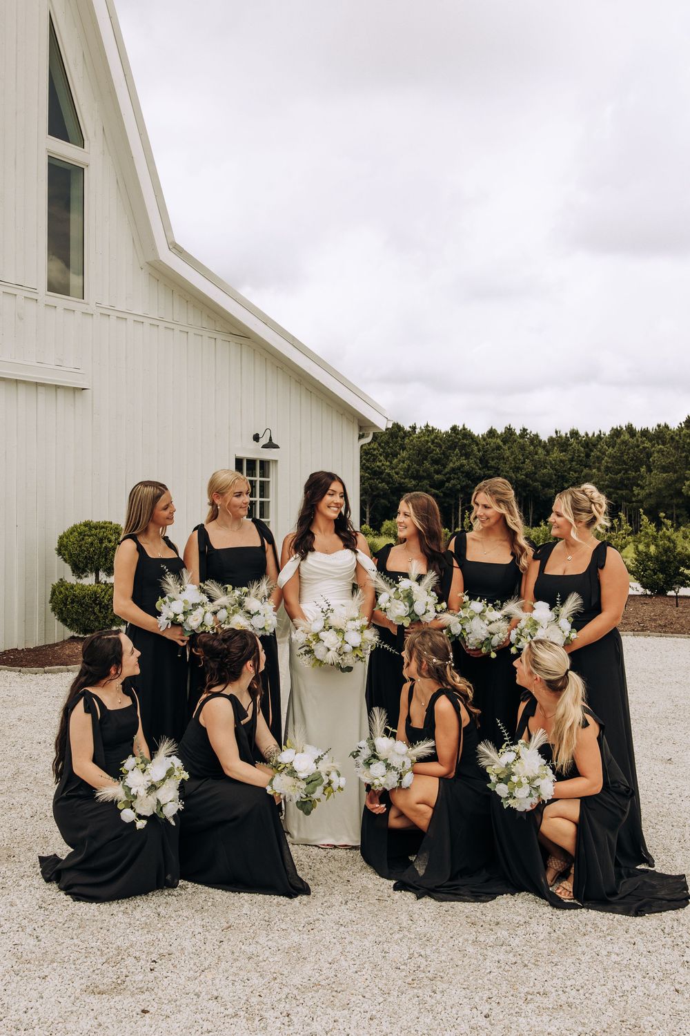 Bride in white gown stands with bridesmaids in black dresses holding white bouquets near a white barn.