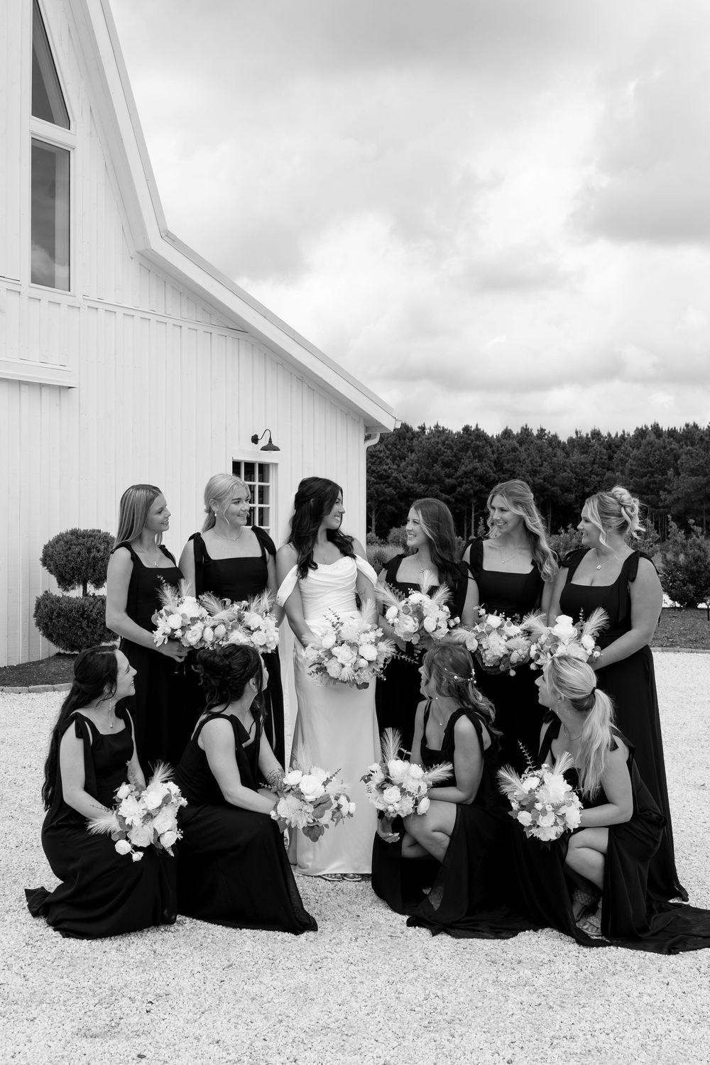 Black and white photo of bride surrounded by bridesmaids in black gowns with white bouquets by a barn.