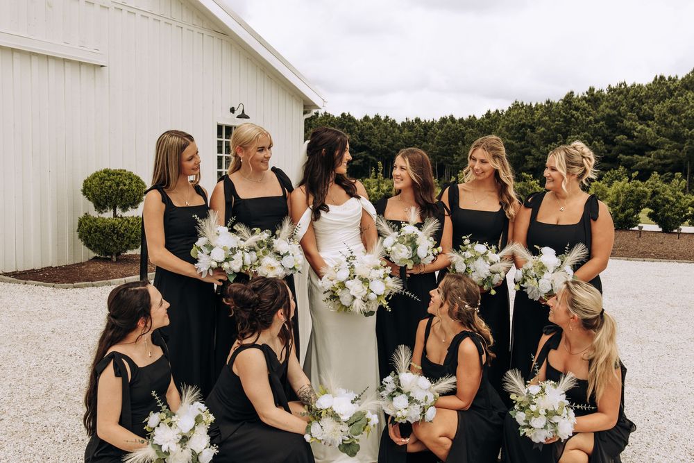 Bride in white dress laughs with bridesmaids in black square-neck gowns holding white floral bouquets.
