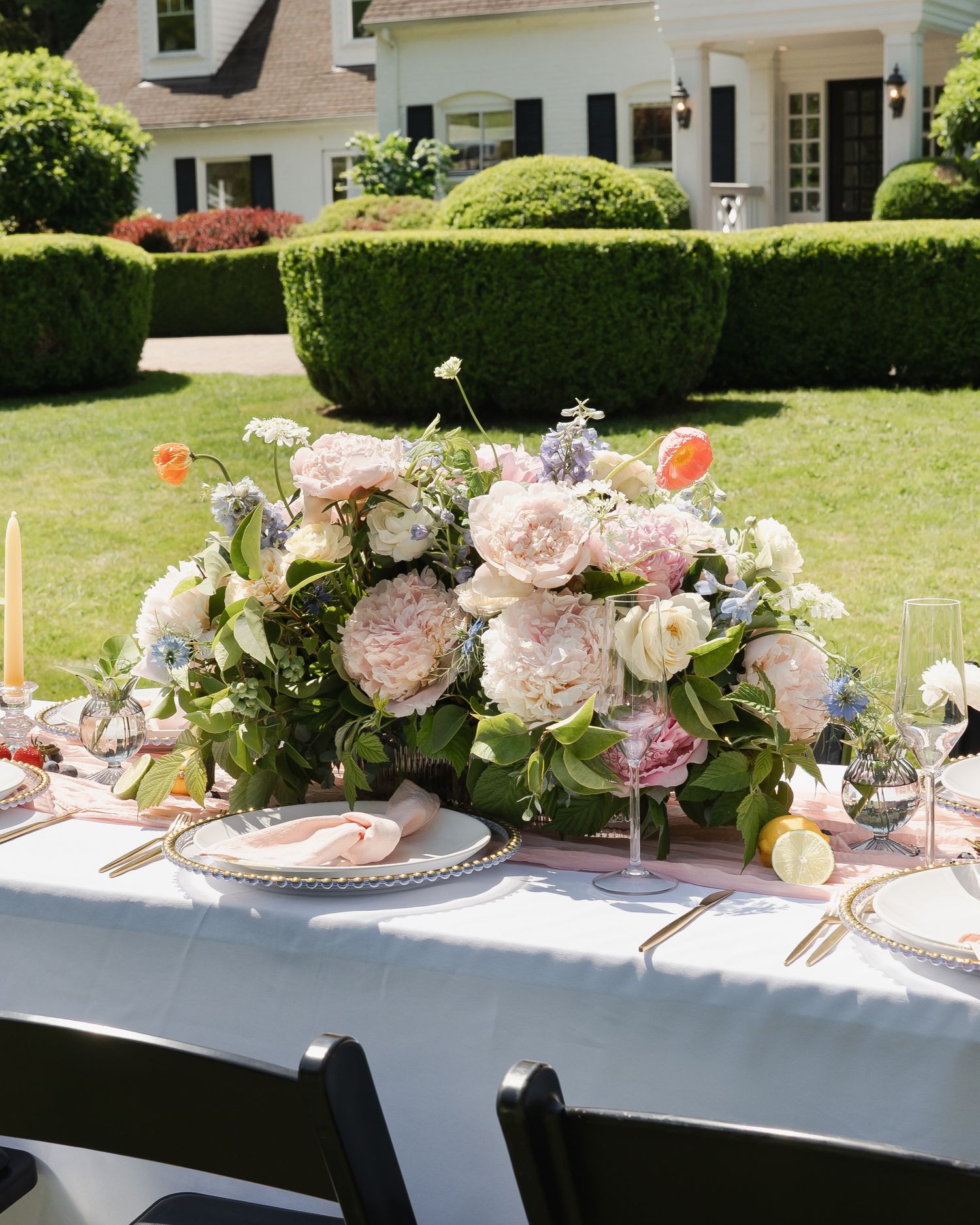 A garden party table setting with pink floral centerpieces and elegant place settings arranged on a white tablecloth outdoors.