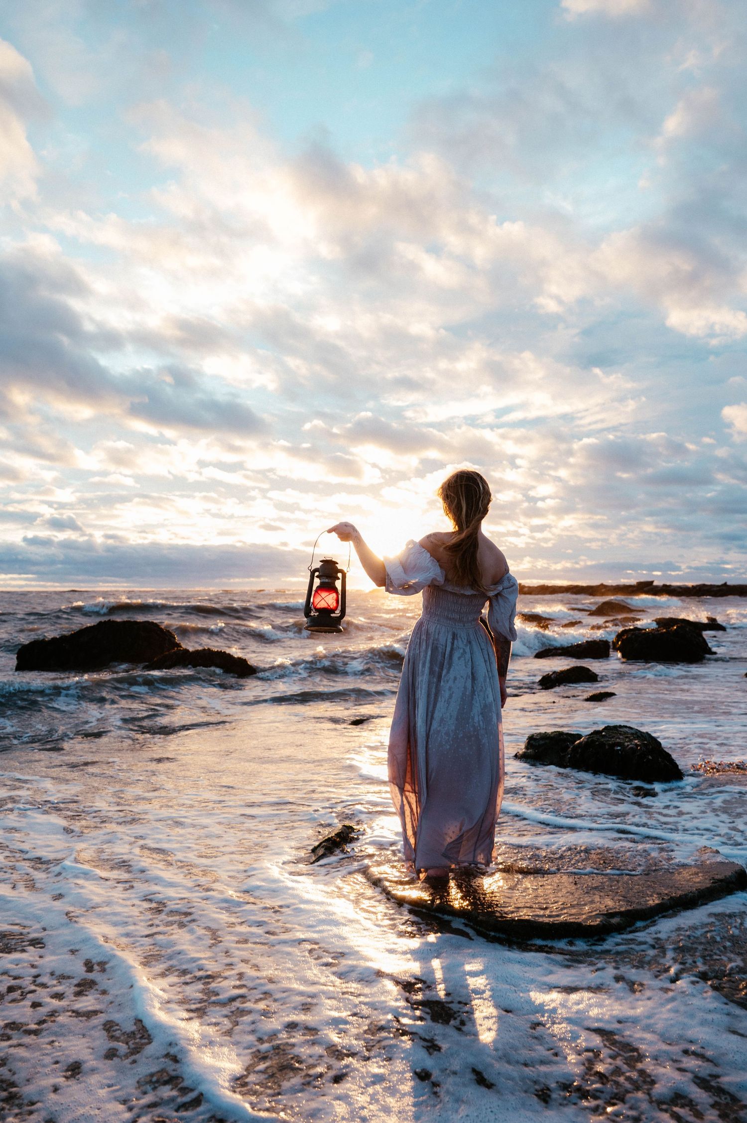 Figure holding lantern on rocky beach at sunset with dramatic cloudy sky.
